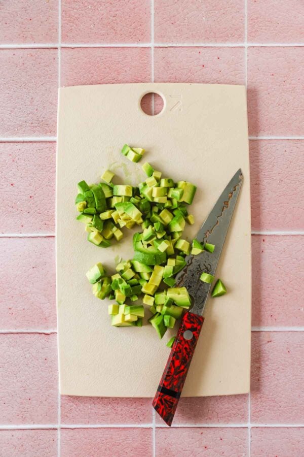 Diced avocado pieces on a white cutting board with a knife, placed on a pink tiled surface.