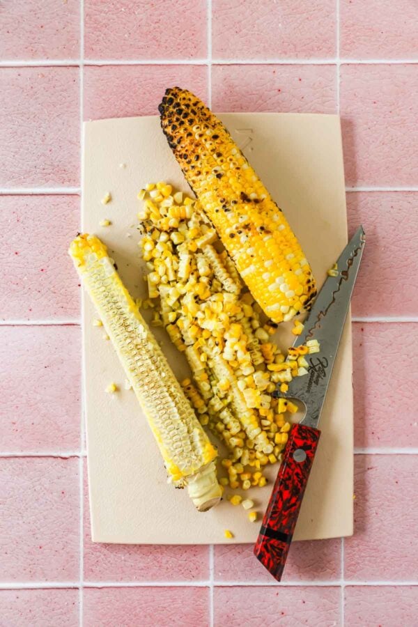 Two grilled corn cobs, one partially cut, lie on a cutting board with a knife and loose corn kernels on a pink tiled surface.