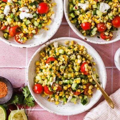 Three bowls of corn salad with cherry tomatoes, cucumber, herbs, and feta sit on a pink tile surface, surrounded by lime, spice, cilantro, and a striped napkin.
