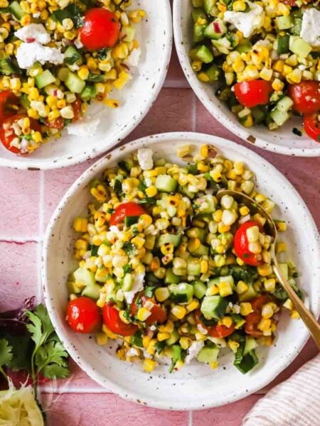 Three bowls of corn salad with cherry tomatoes, cucumber, herbs, and feta sit on a pink tile surface, surrounded by lime, spice, cilantro, and a striped napkin.