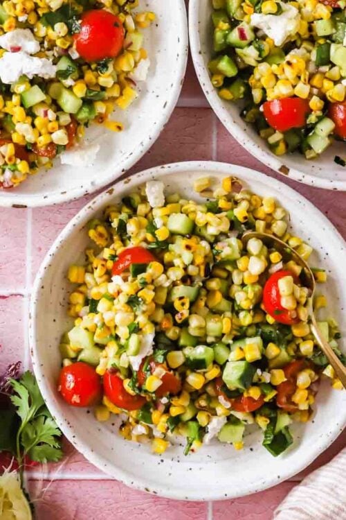 Three bowls of corn salad with cherry tomatoes, cucumber, herbs, and feta sit on a pink tile surface, surrounded by lime, spice, cilantro, and a striped napkin.