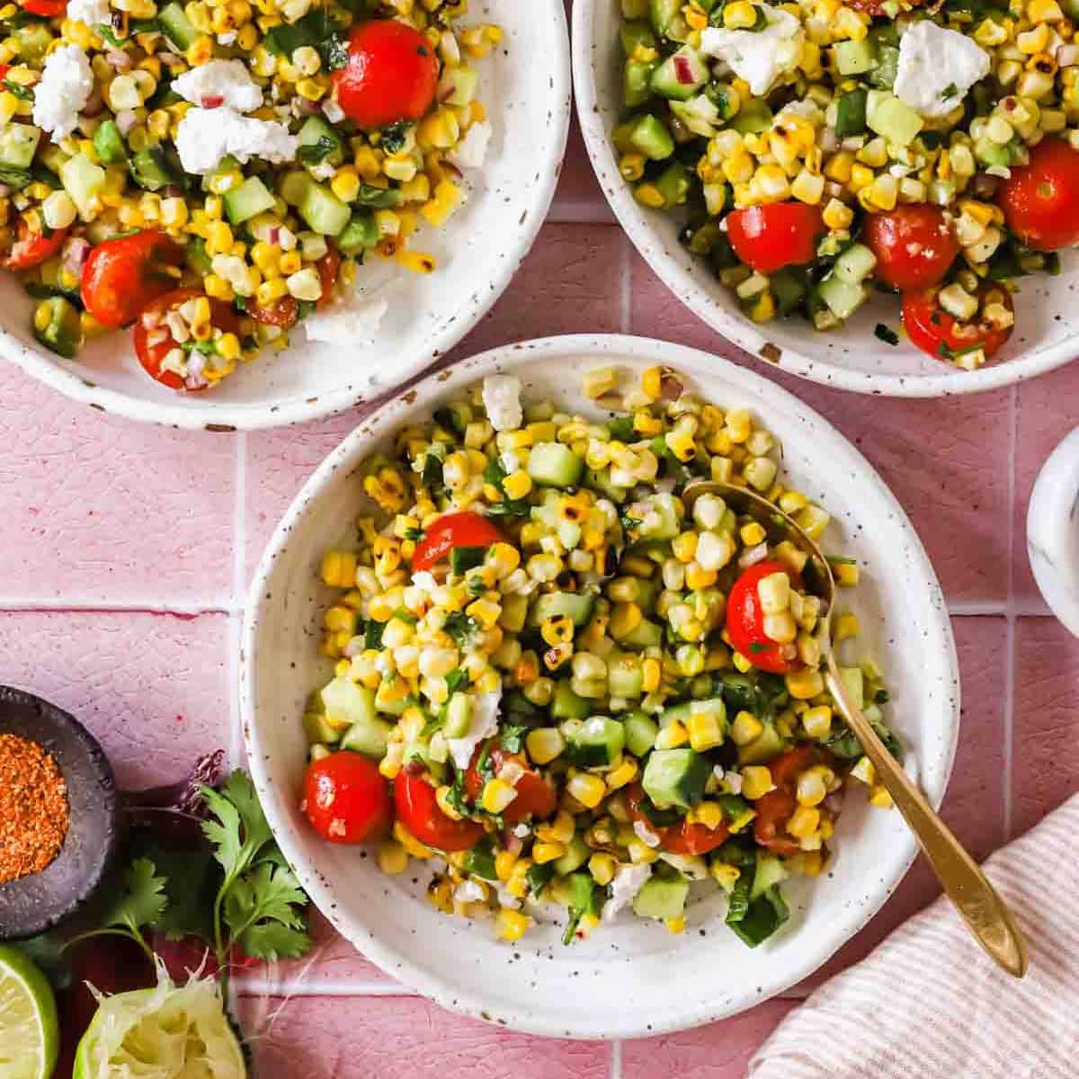 Three bowls of corn salad with cherry tomatoes, cucumber, herbs, and feta sit on a pink tile surface, surrounded by lime, spice, cilantro, and a striped napkin.