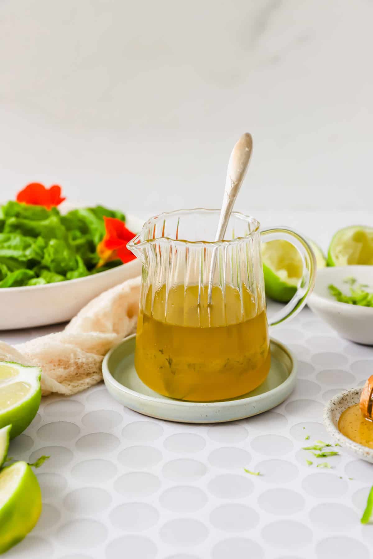 A glass pitcher with a silver spoon contains yellow salad dressing, placed on a small plate. Fresh limes, lettuce, and a bowl are visible in the background.