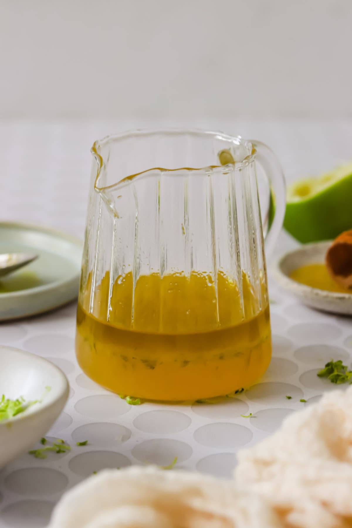 A glass pitcher of yellow salad dressing with a spoon inside, surrounded by limes, a bowl of greens, shredded zest, and honey on a white patterned surface.