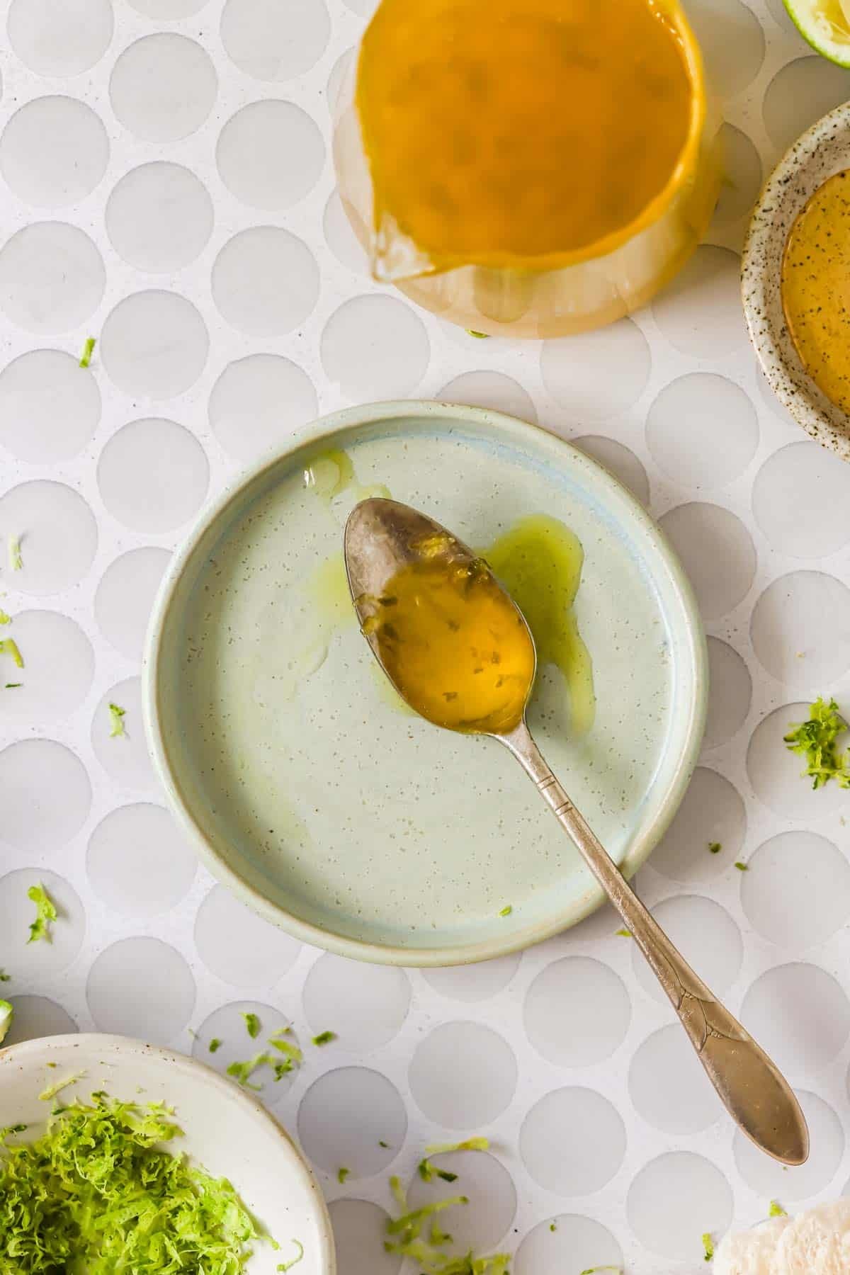 A spoon with golden liquid, likely honey, rests on a small round plate. Surrounding the plate are bowls and scattered green herb pieces on a white patterned surface.