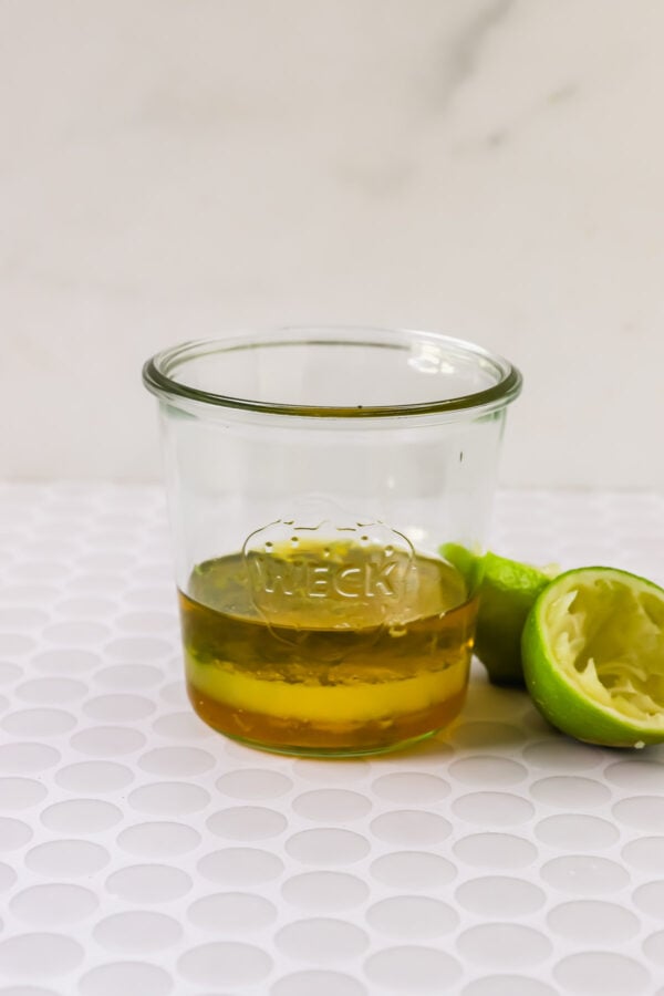 A glass jar with a small amount of golden liquid sits on a white surface next to a squeezed lime half.