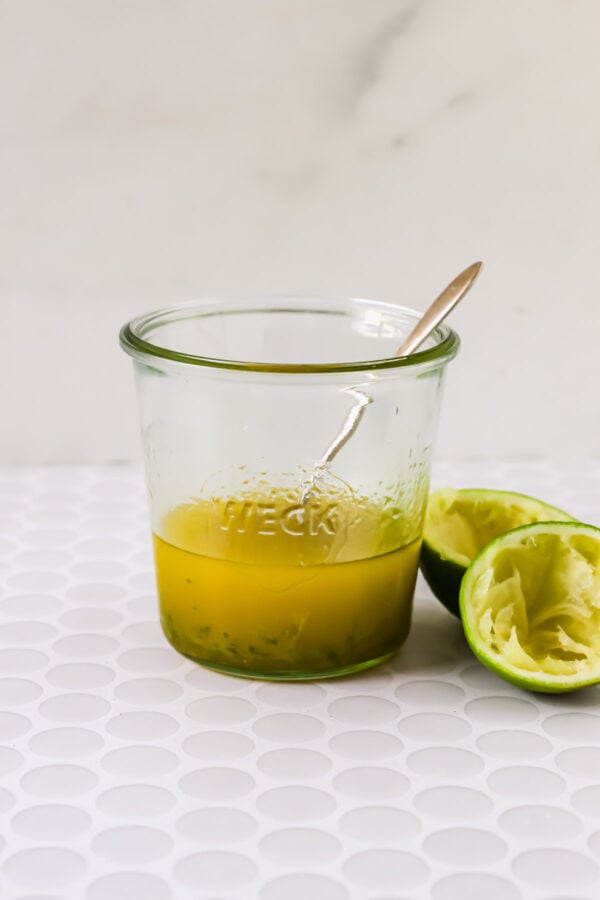A glass jar with yellow liquid and a spoon inside sits next to two squeezed lime halves on a white textured surface.