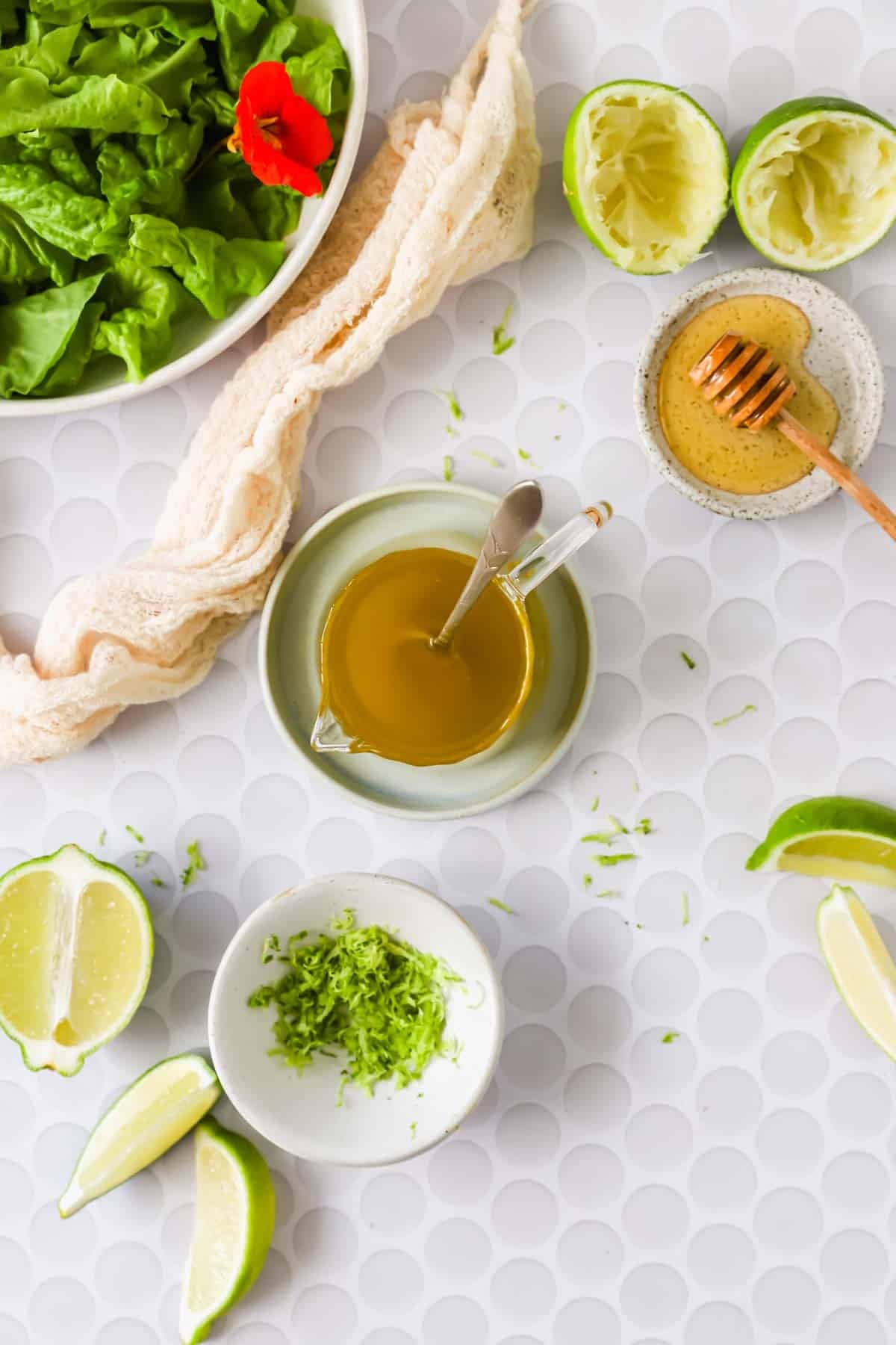 A bowl of lettuce, squeezed limes, a dish of honey, a bowl of olive oil with a spoon, and a plate of lime zest are arranged on a white surface.