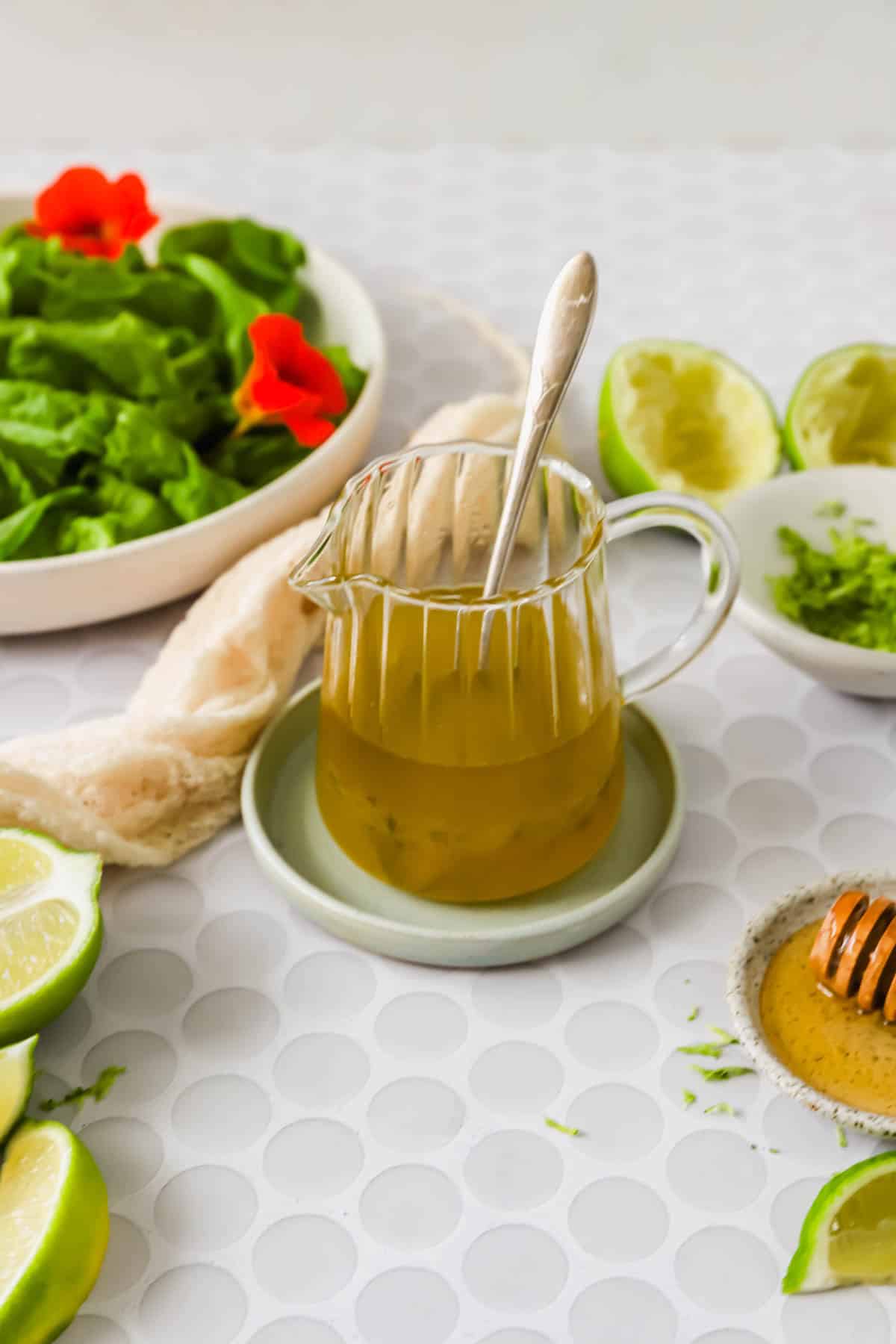 A glass pitcher of salad dressing with a spoon, surrounded by lime slices, lettuce, a honey dipper, and a bowl of zest on a white patterned surface.