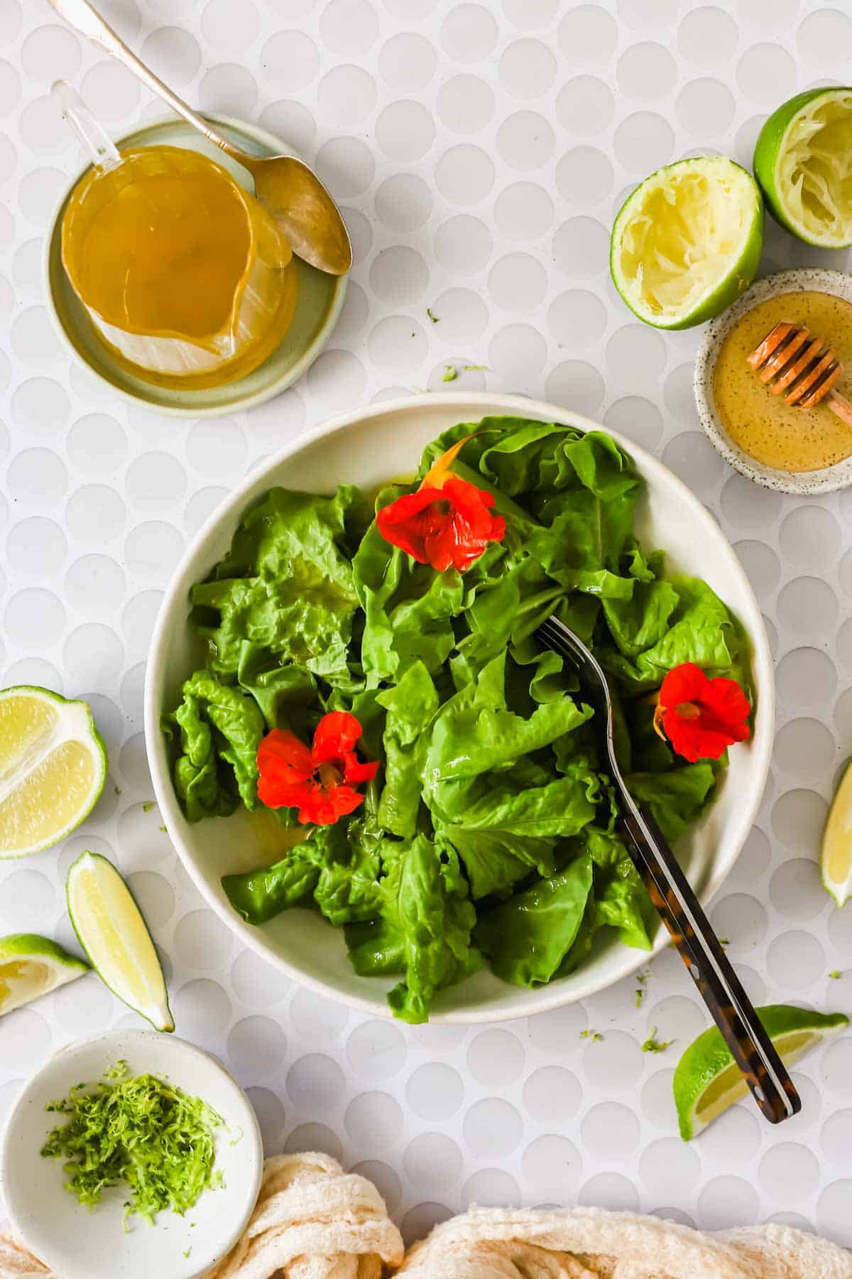 Bowl of green lettuce topped with red edible flowers, surrounded by lime wedges, honey, dressing, and a grater on a white patterned surface.