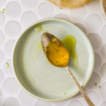 A spoon with honey rests on an empty plate atop a white patterned surface, with lime zest scattered nearby.