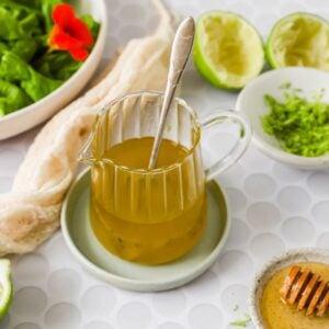 A glass pitcher of yellow salad dressing with a spoon inside, surrounded by limes, a bowl of greens, shredded zest, and honey on a white patterned surface.