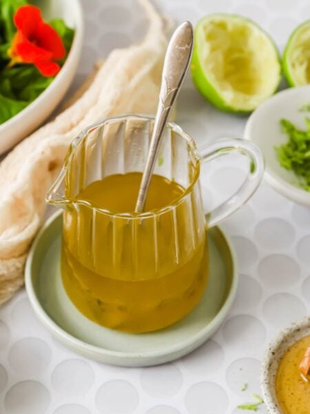 A glass pitcher of yellow salad dressing with a spoon inside, surrounded by limes, a bowl of greens, shredded zest, and honey on a white patterned surface.