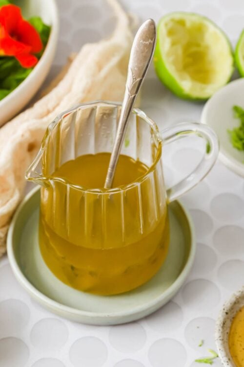 A glass pitcher of yellow salad dressing with a spoon inside, surrounded by limes, a bowl of greens, shredded zest, and honey on a white patterned surface.