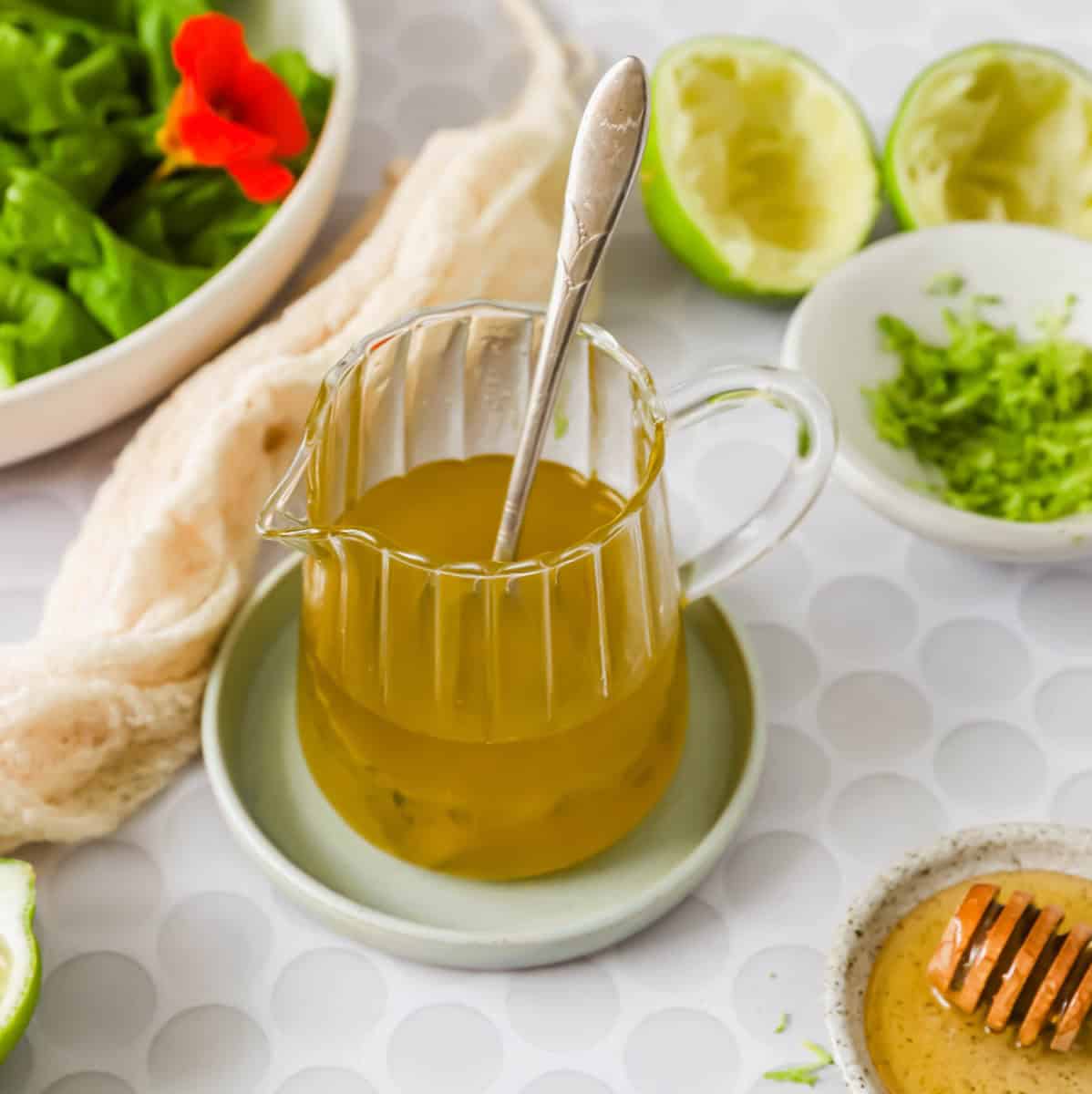 A glass pitcher of yellow salad dressing with a spoon inside, surrounded by limes, a bowl of greens, shredded zest, and honey on a white patterned surface.