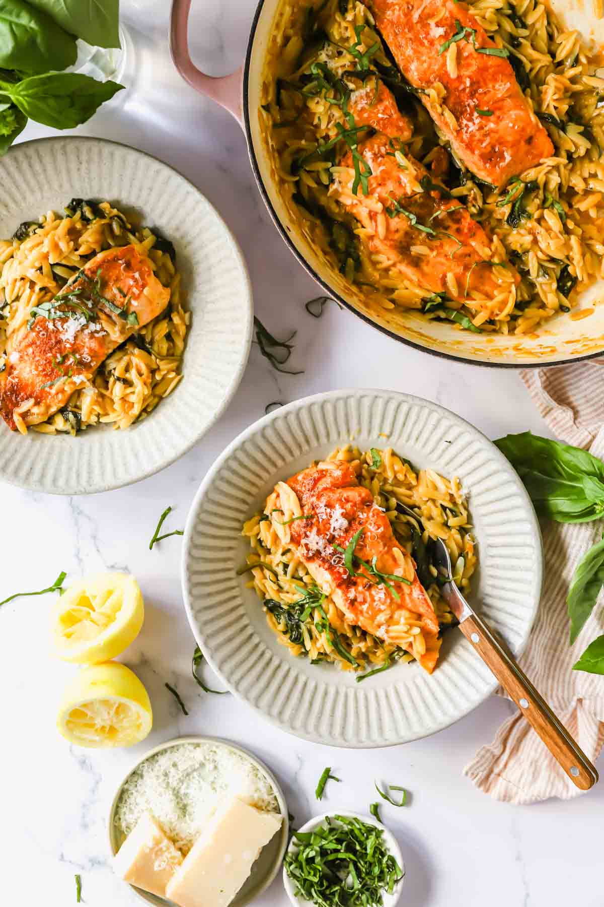 Salmon fillets served over orzo pasta with spinach in a creamy sauce, plated in bowls with lemon halves, basil, and Parmesan cheese on the side.