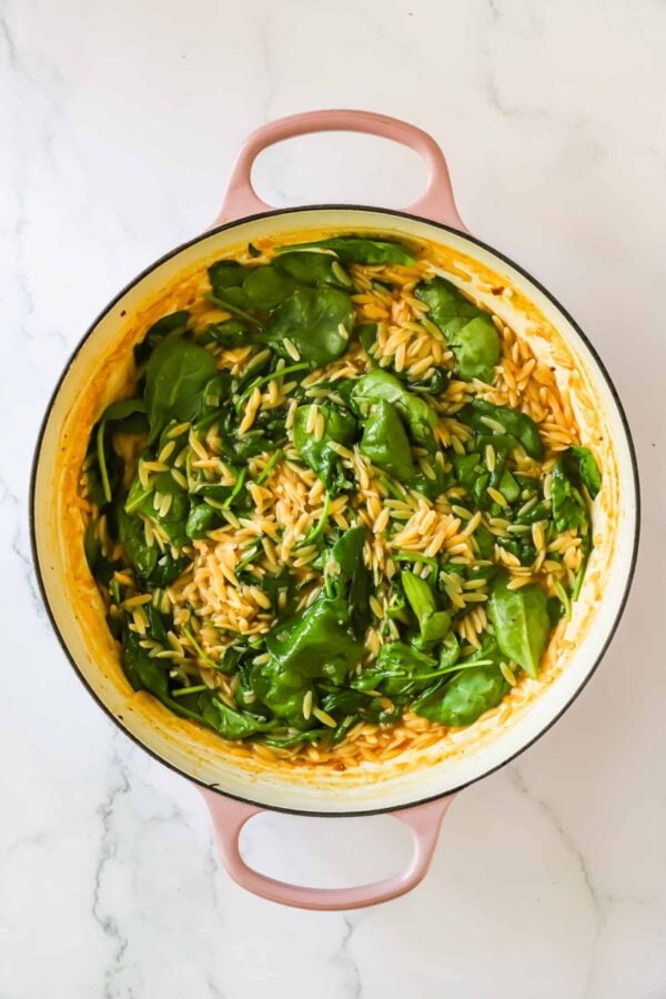 A pink pot filled with cooked orzo pasta mixed with fresh spinach, placed on a white marble surface.