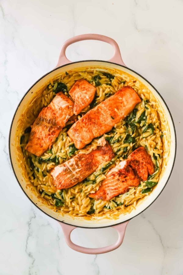 Four cooked salmon fillets on a bed of creamy orzo pasta with spinach in a round, light-colored pot on a white marble surface.
