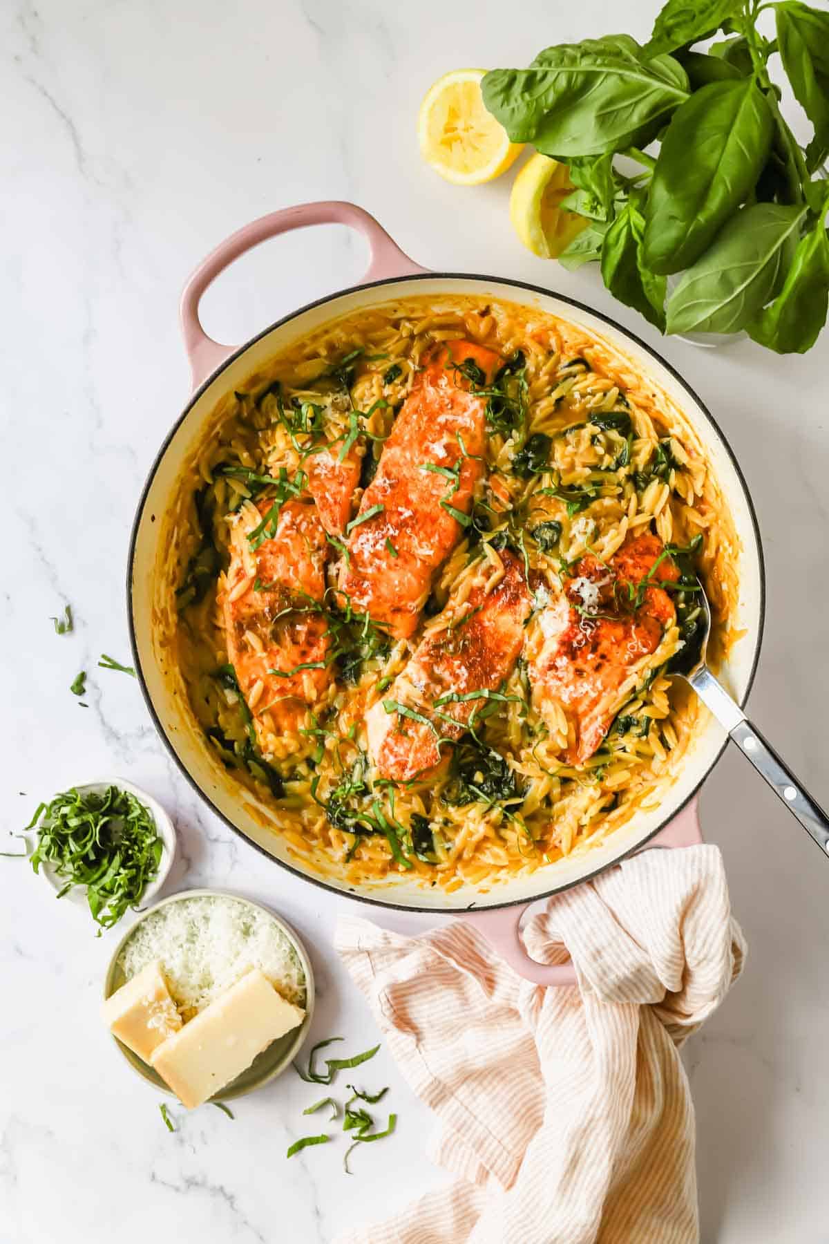 A skillet with cooked salmon fillets on creamy orzo pasta, garnished with herbs, next to fresh basil, grated cheese, and a lemon half on a marble surface.