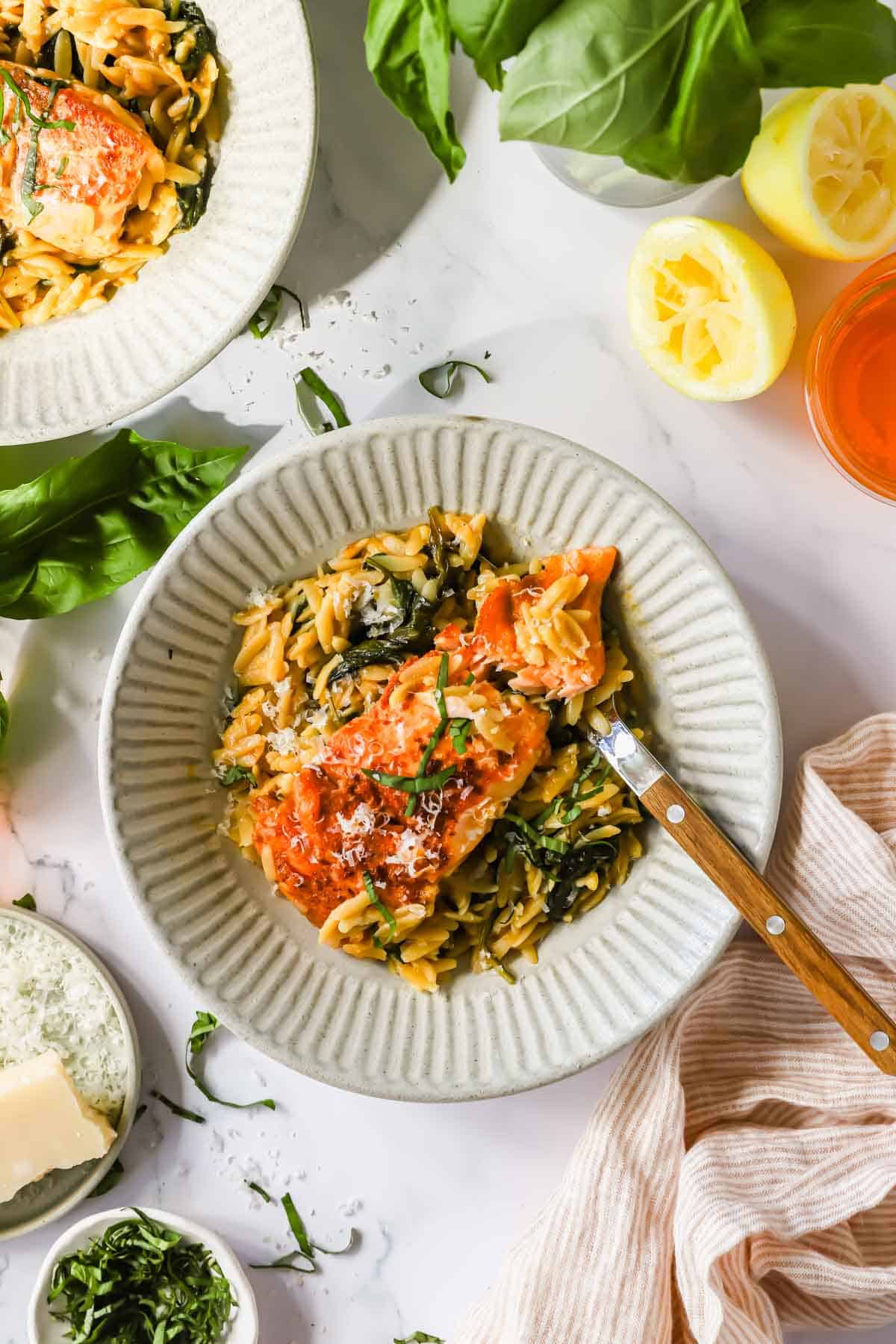 A bowl of orzo pasta with spinach, topped with a piece of salmon and grated cheese, with a fork, fresh basil, lemon, and a glass of orange drink nearby.