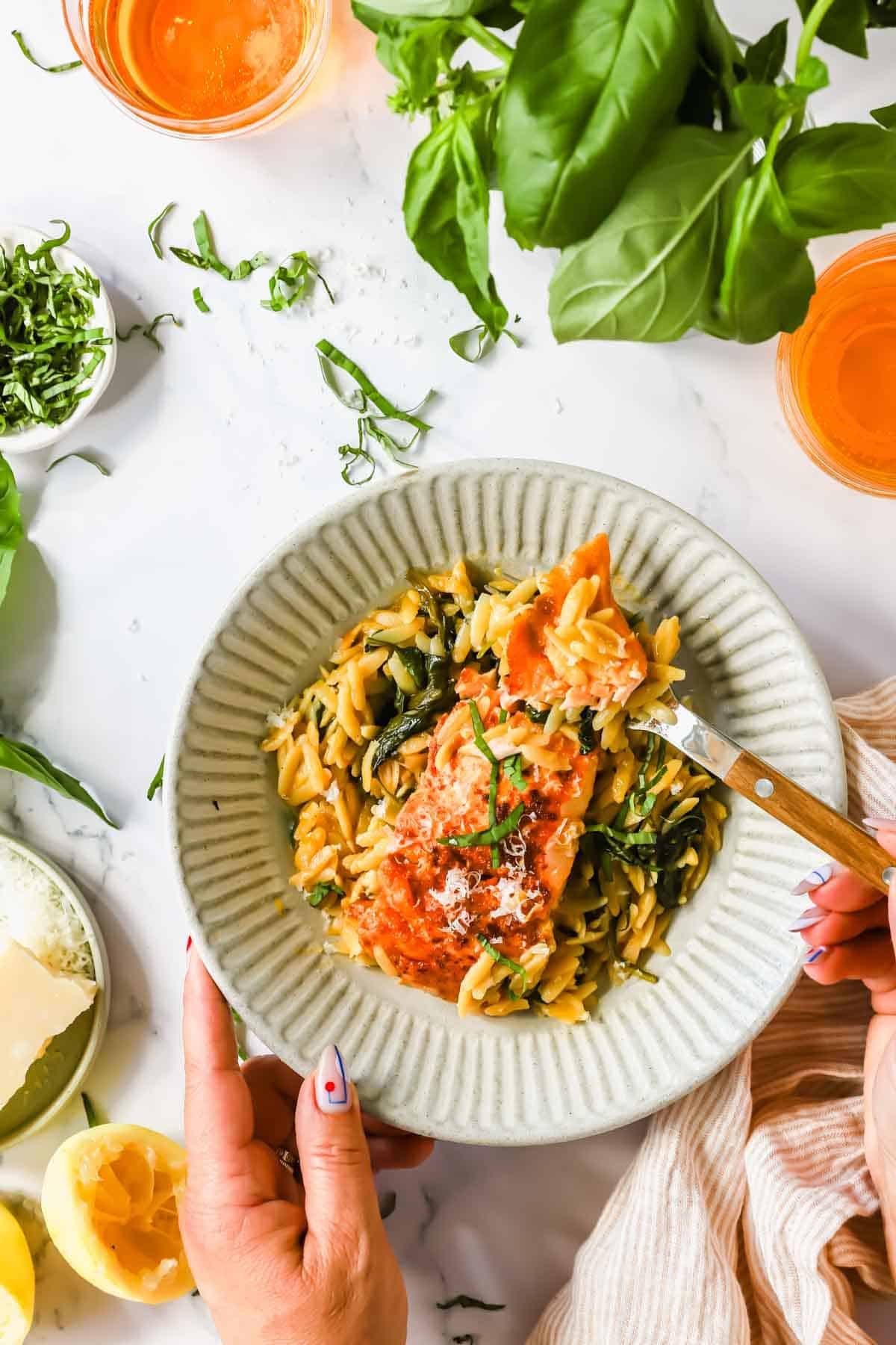 A person holds a bowl of orzo pasta with spinach and tomato, garnished with herbs and cheese, next to lemon, basil, and drinks on a white table.