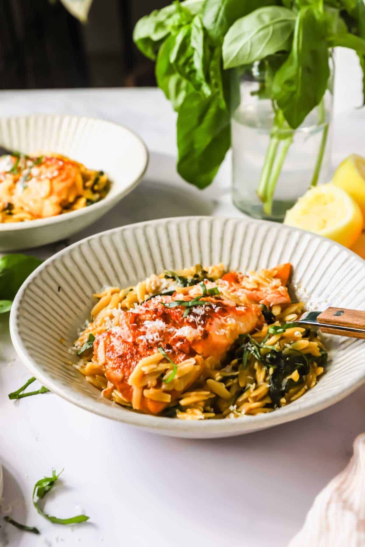 A bowl of orzo pasta with tomato sauce, spinach, and grated cheese, garnished with fresh basil. A lemon wedge and basil plant are in the background.