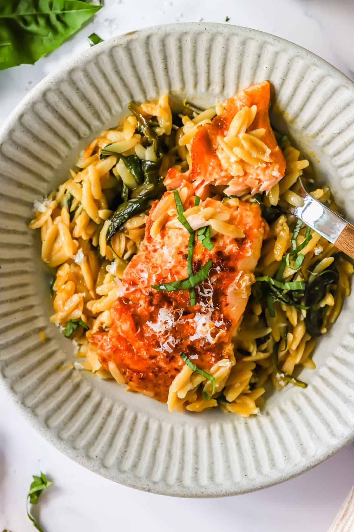 A plate of orzo pasta with wilted greens and a piece of salmon, topped with grated cheese and chopped fresh herbs.