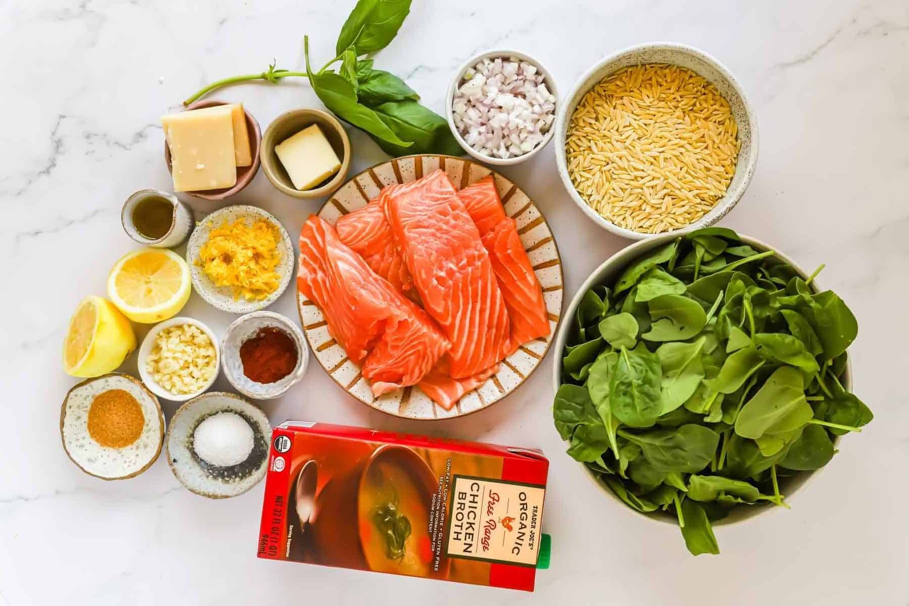 Top-down view of ingredients on a white surface: fresh spinach, orzo, shallots, basil, butter, garlic, cheese, lemon, spices, salmon fillets, and a box of organic chicken broth.