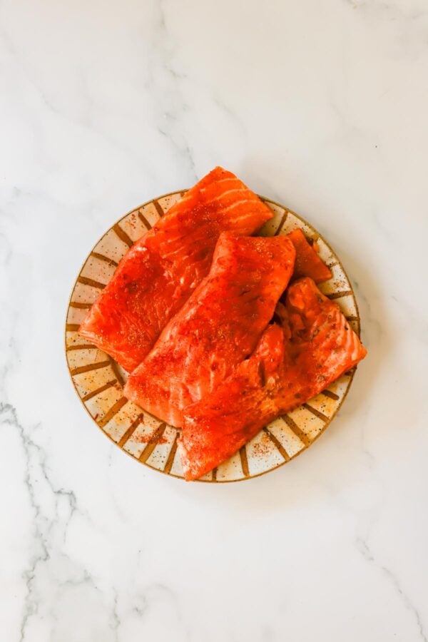 Three raw salmon fillets with seasoning are arranged on a striped ceramic plate, placed on a white marble surface.