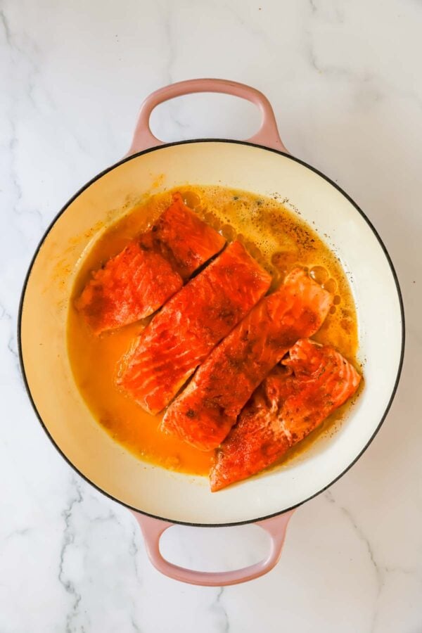 Four cooked salmon fillets sit in a round, cream-colored pan with orange sauce, placed on a white marble surface.