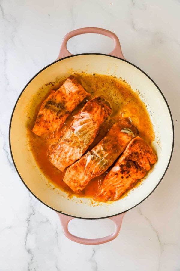 Three cooked salmon fillets in sauce sit inside a round, light-colored pan with pink handles on a white marble surface.