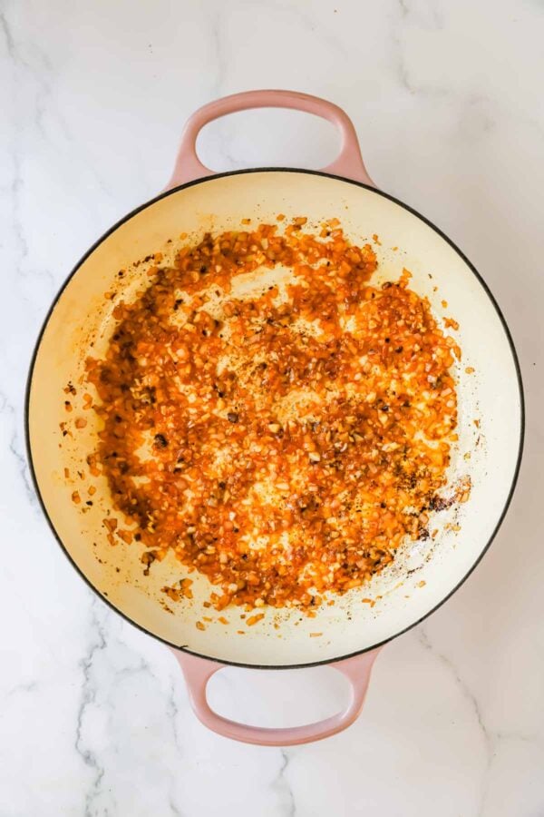 Diced onions and carrots sautéing with tomato paste and seasonings in a large, round, light-colored pot on a marble surface.
