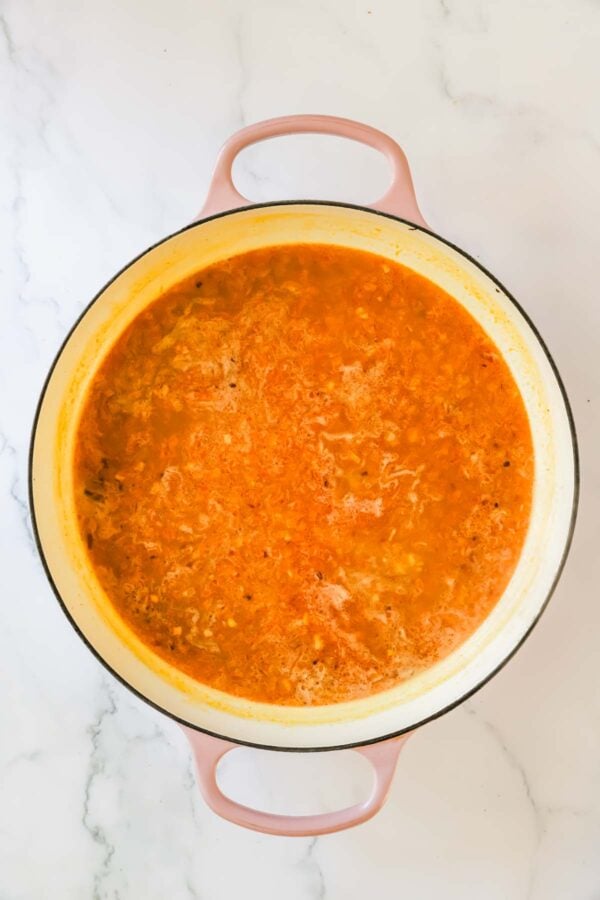 A large pink-handled pot filled with simmering orange-red soup or stew sits on a white marble surface.