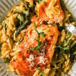 A plate of cooked salmon fillets served over orzo pasta with spinach, garnished with herbs and grated cheese.
