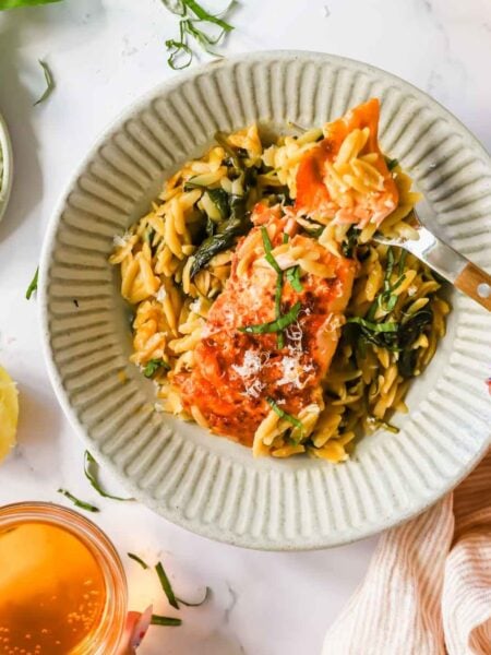 A plate of orzo pasta with cooked salmon and greens, garnished with herbs, next to a drink, lemon half, and a small dish of grated cheese.