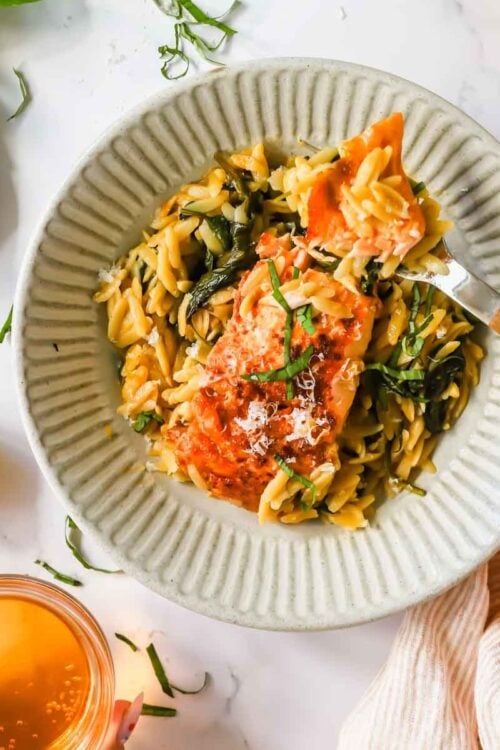 A plate of orzo pasta with cooked salmon and greens, garnished with herbs, next to a drink, lemon half, and a small dish of grated cheese.