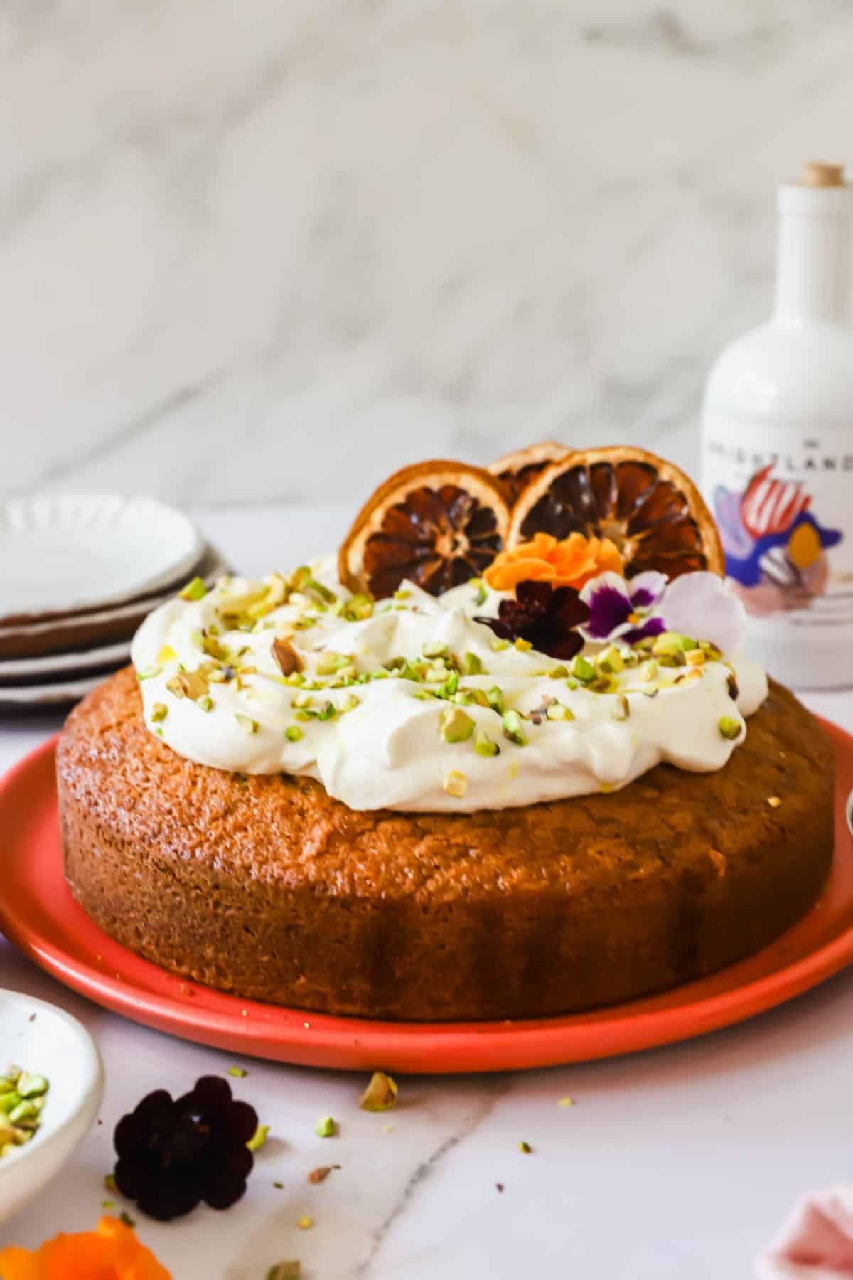 A round cake topped with whipped cream, chopped pistachios, dried orange slices, and edible flowers sits on a red plate.