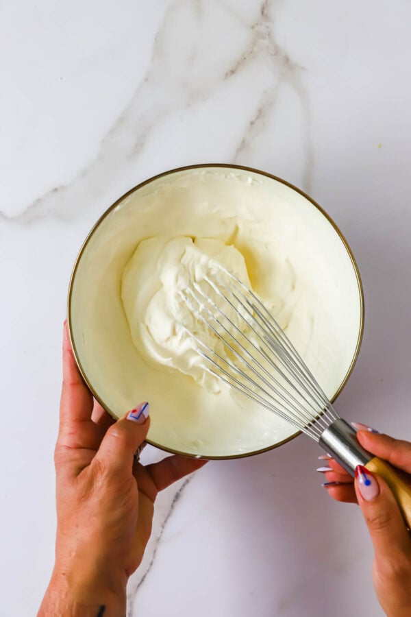 A person whisking thick white cream in a white bowl on a marble surface.