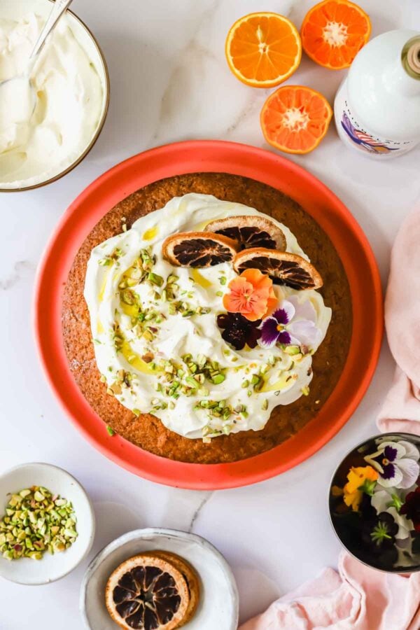 A round cake with white frosting, topped with dried orange slices, chopped pistachios, and edible flowers, on a red plate surrounded by ingredients on a white surface.