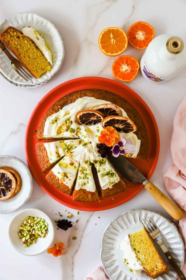 A round orange cake with white frosting, dried citrus slices, and chopped pistachios, surrounded by plates with cake slices, orange halves, and a bottle on a white surface.