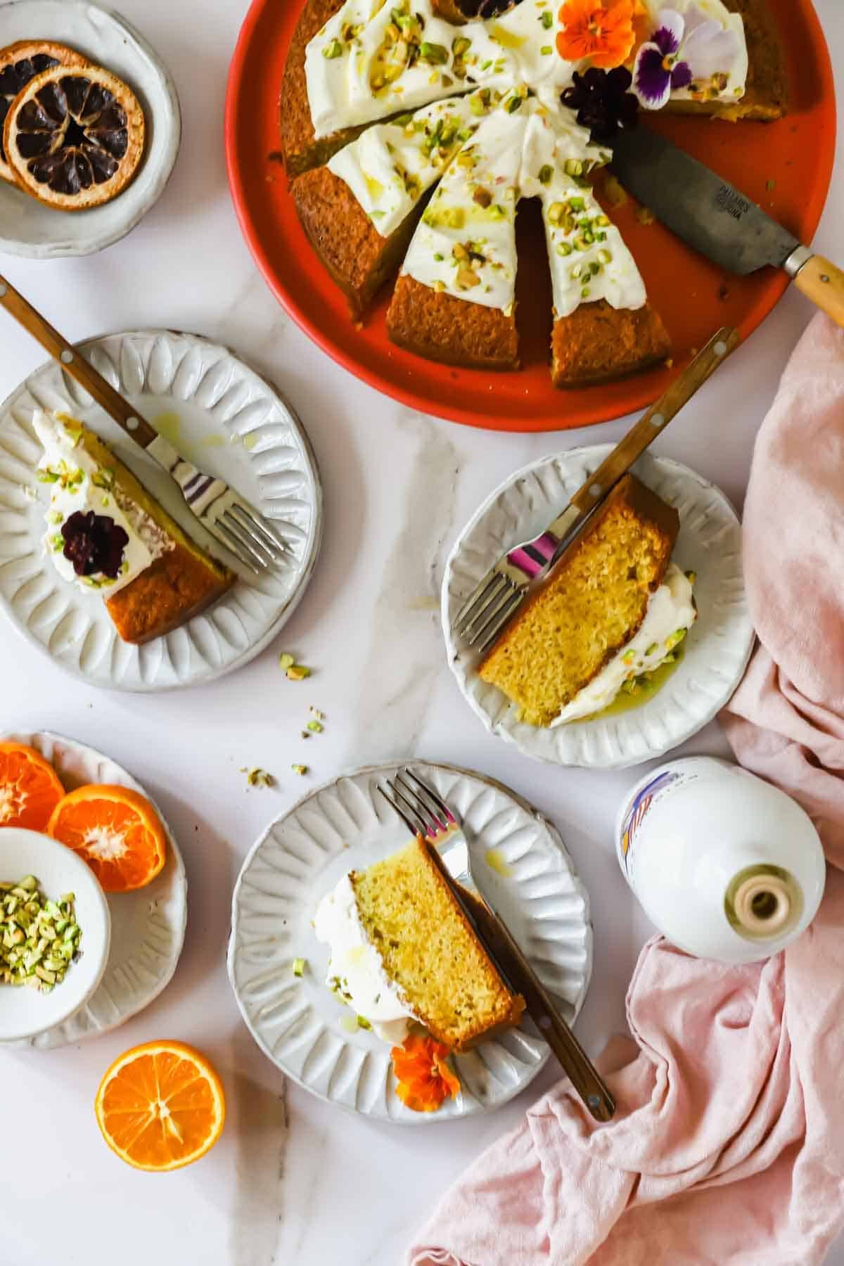 A partially sliced cake with white frosting and flower garnish is served on plates with forks, alongside orange slices, chopped pistachios, and a bottle on a white table.