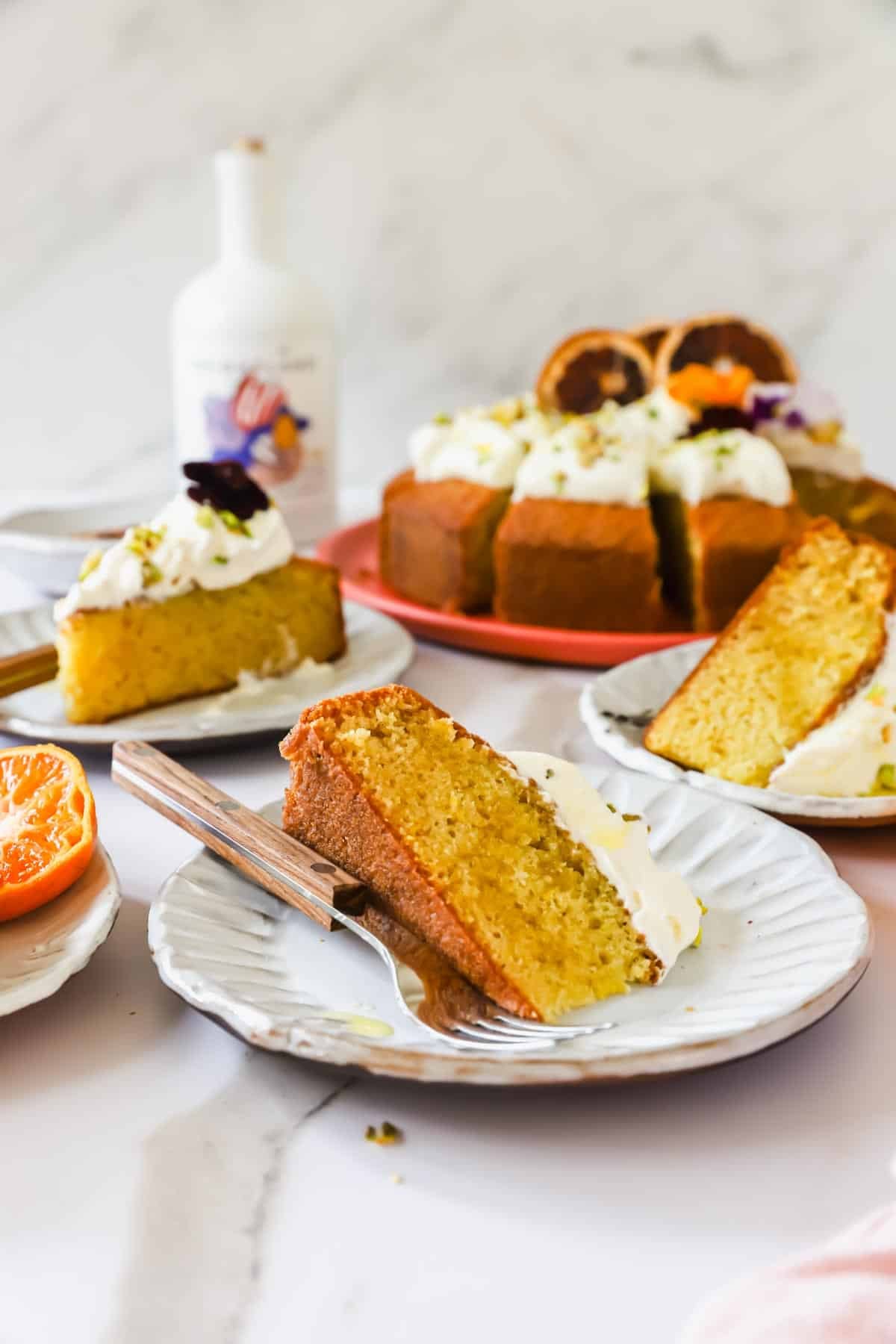 Slices of orange cake with whipped cream on plates, garnished with chopped pistachios. A full cake with cream and dried orange on a red plate is in the background.