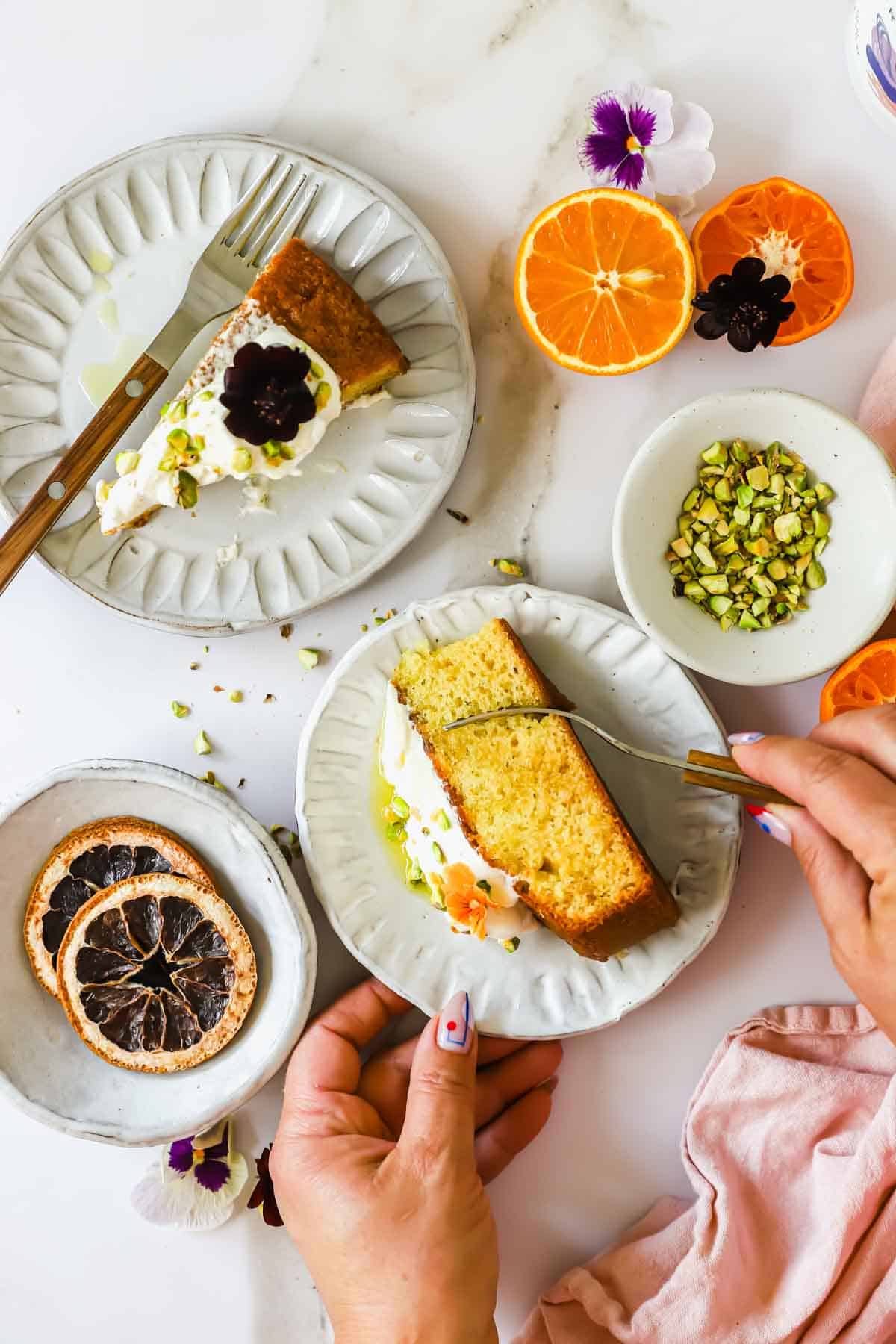 A person slices a piece of cake topped with cream and chopped pistachios on a white plate, surrounded by citrus fruits, flower garnishes, and a fork.