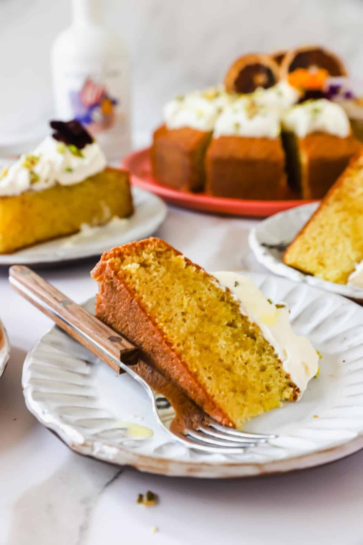 A slice of yellow cake with white frosting on a plate with a fork; more cake slices and a whole cake are in the background.