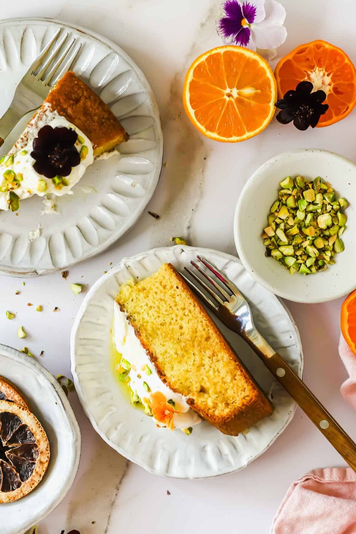 A slice of yellow cake with cream and pistachios on a white plate, a fork, orange halves, flower garnish, and a small bowl of chopped pistachios nearby.