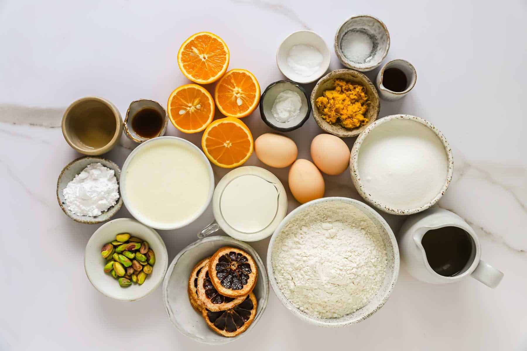 Assorted baking ingredients including flour, sugar, eggs, cream, oranges, pistachios, dried fruits, vanilla, baking powder, and measuring cups arranged neatly on a white surface.