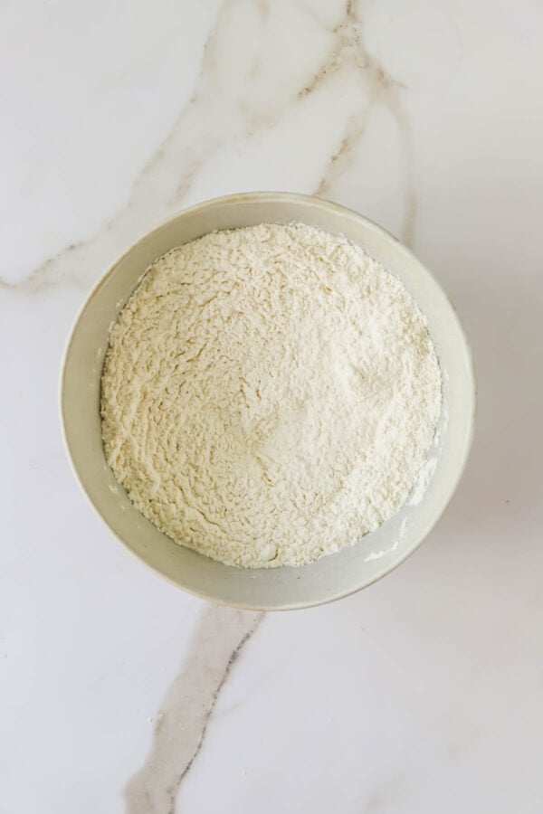 A white bowl filled with flour sits on a white marble surface.