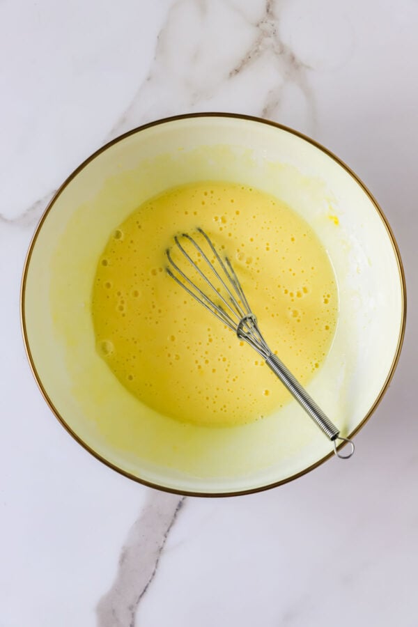 A metal whisk rests in a mixing bowl filled with a frothy yellow liquid, likely a batter or egg mixture, on a white marble surface.