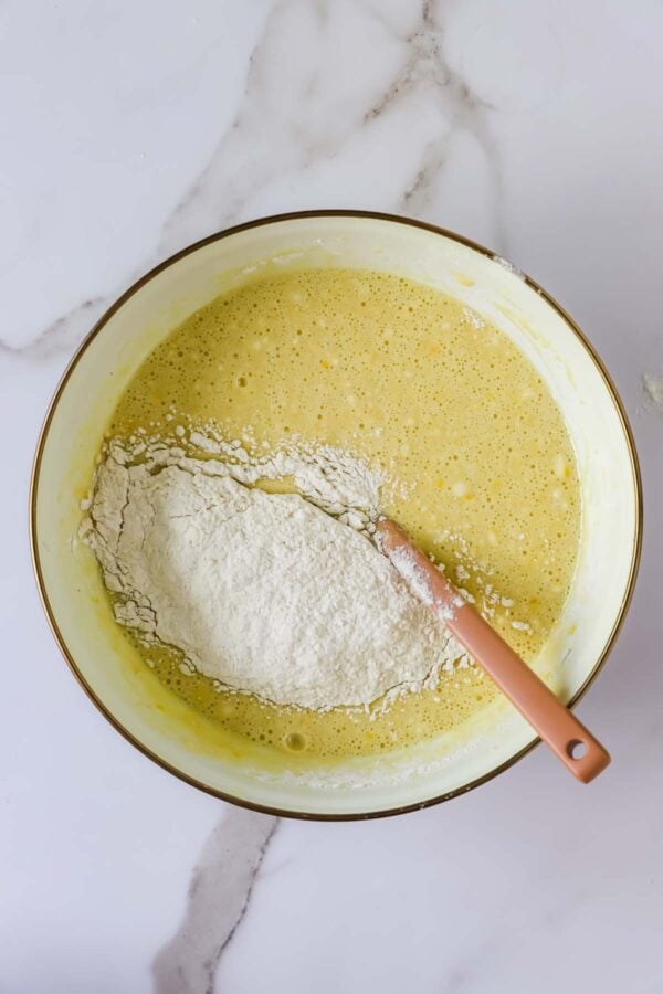 A mixing bowl with a yellow batter and a pile of flour on top, being stirred with a pink spatula on a marble countertop.