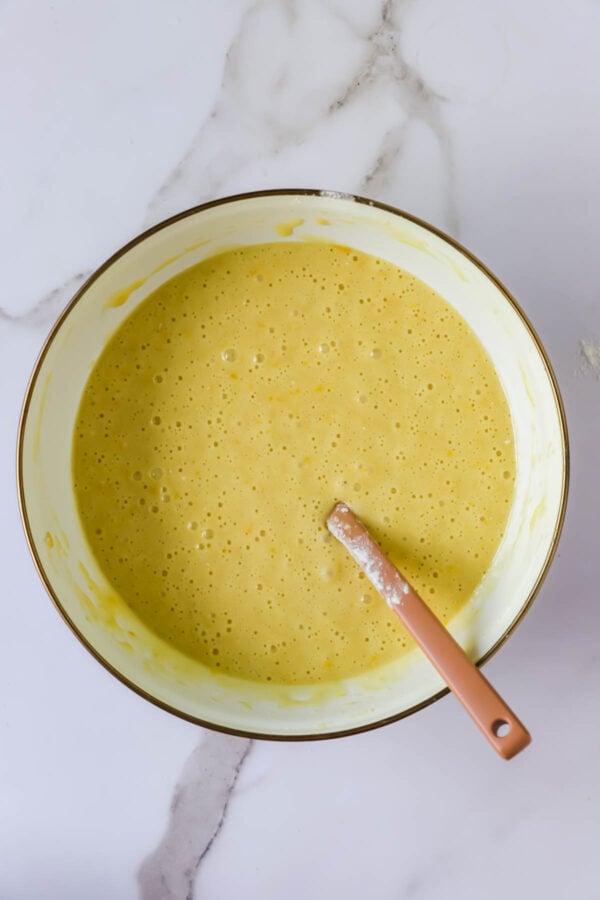 A mixing bowl filled with yellow batter and a spatula resting inside, placed on a white marble surface.